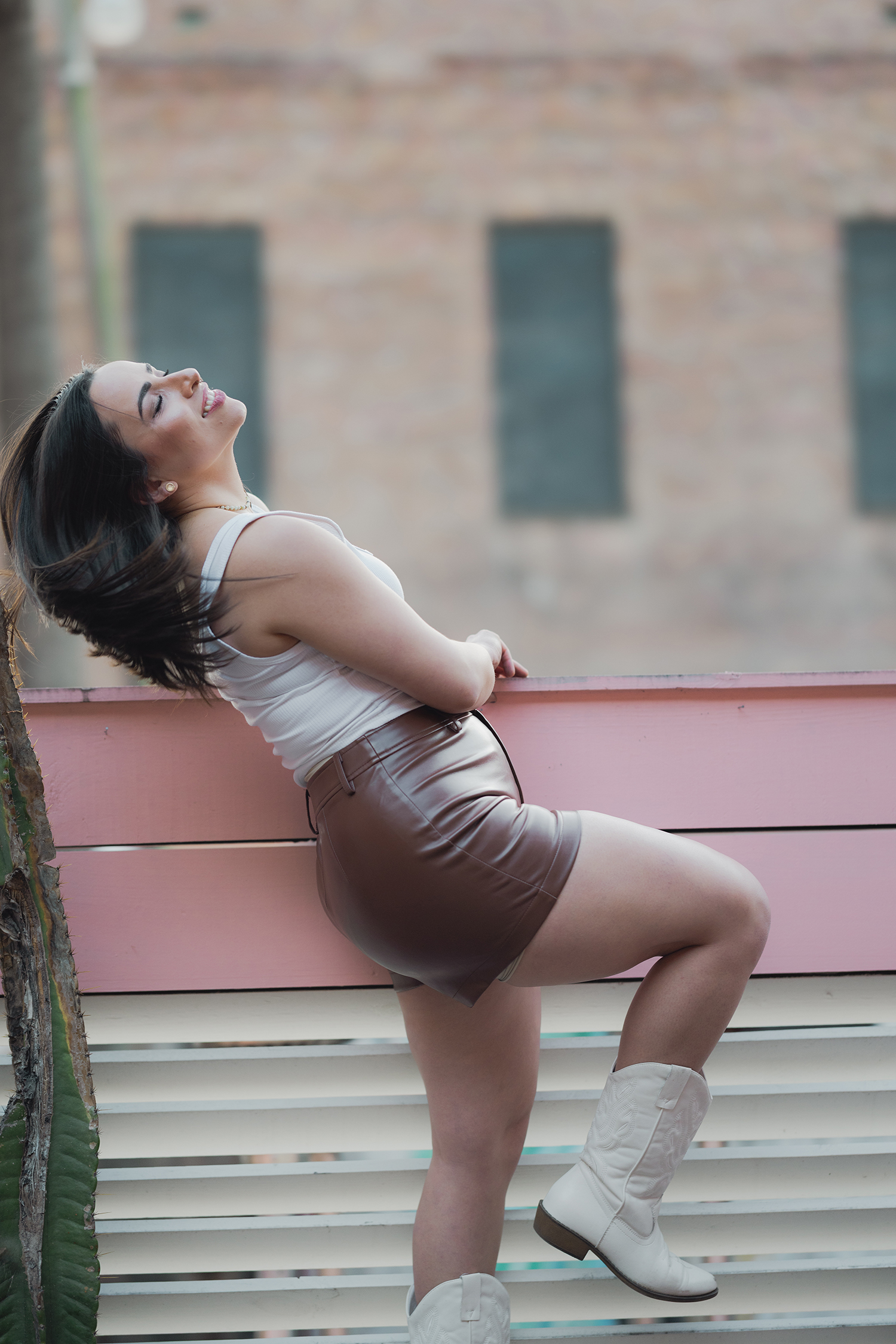 A woman wearing a white tank top and leather shorts stands on a railing and shakes her head, smiling
